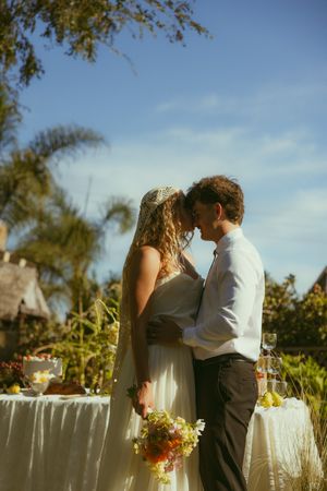 Bride and groom share a romantic kiss outdoors at a sunny garden wedding ceremony with tropical palms.