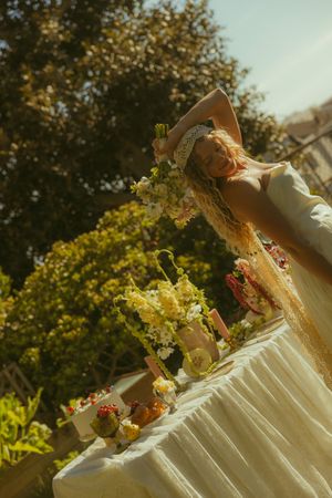 Blonde woman in white dress and hat poses by floral table outdoors in sunny garden setting.