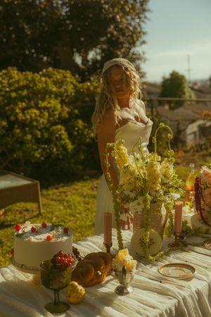 Bride in white dress arranging yellow flowers on an outdoor table with cake and candles in golden sunlight.