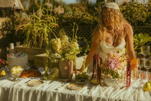 Bride in white dress arranging floral centerpieces on an elegant outdoor table with lush greenery in background.