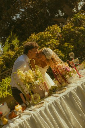 Couple sharing a kiss at an outdoor wedding reception table adorned with lush floral arrangements in a sunlit garden.