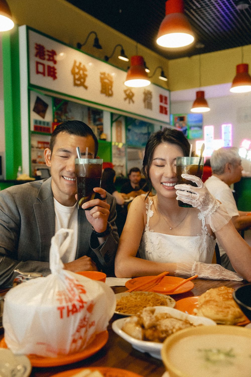 A smiling Asian couple in wedding attire toasting drinks at a casual Asian restaurant with Chinese signage and food on the table.