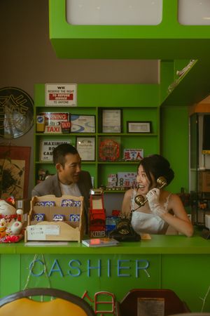 Couple smiling at a green cashier counter, woman holding vintage telephone, surrounded by retro decor and signs.