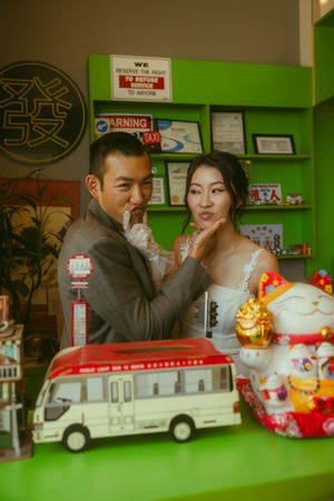 Asian couple posing romantically in a colorful shop with a lucky cat figurine and toy bus on display.
