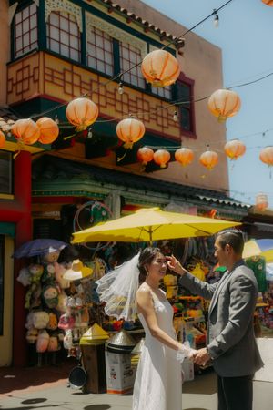 Bride and groom hold hands in Chinatown, surrounded by orange lanterns and colorful storefronts on a sunny day.