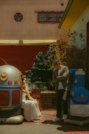 Bride in white dress sits on coin-operated robot ride while groom stands beside her at colorful amusement area.