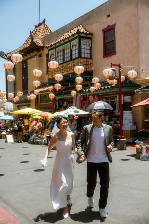 Newlywed Asian couple holding hands walking through Chinatown with paper lanterns and colorful buildings.