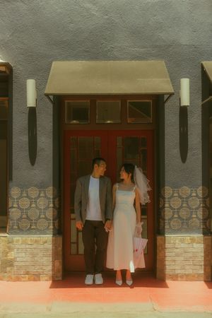 A couple in wedding attire stands holding hands before a red door with tiled walls and wall sconces.