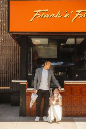 Couple posing outside Frank n Fr restaurant, man in gray blazer standing, woman in white dress sitting by orange storefront.