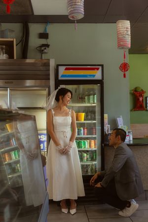 Bride in white dress and veil smiles at groom kneeling before her inside a cozy Asian restaurant.