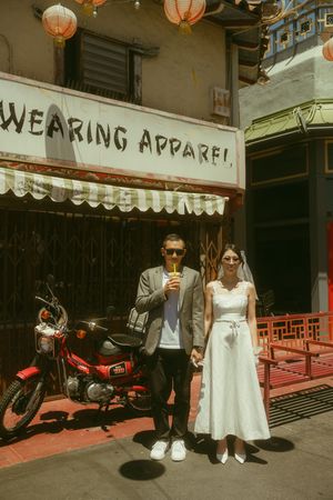 Couple in wedding attire posing outside a Chinatown apparel shop with red lanterns and a vintage motorcycle.