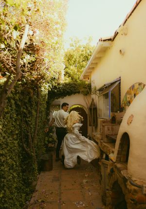 A couple in wedding attire dancing in a sunlit courtyard between lush greenery and a yellow stucco building.