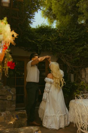 A bride in a white off-shoulder gown dances with a groom outdoors, surrounded by lush greenery and colorful decorations.