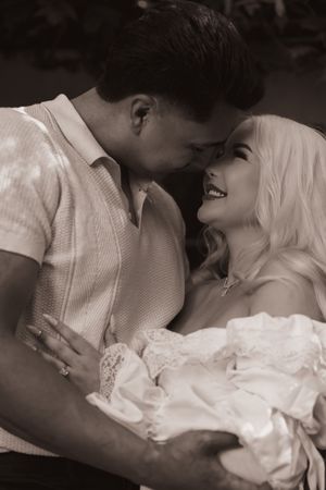 Black and white photo of a romantic couple sharing an intimate moment while holding a newborn baby.