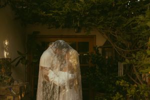 Bride covered in lace veil standing among lush green trees and plants in a garden setting.