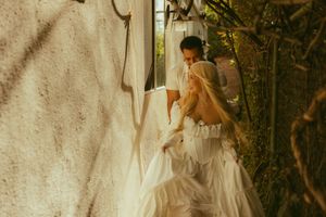 Bride in white gown with veil assisted by bridesmaid near rustic wall and lush greenery.