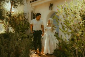 Couple holding hands walking along a garden path outside a white Spanish-style home in warm sunlight.