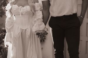 Black and white photo of a bride in a white corset gown holding a bouquet beside a groom in dark pants.
