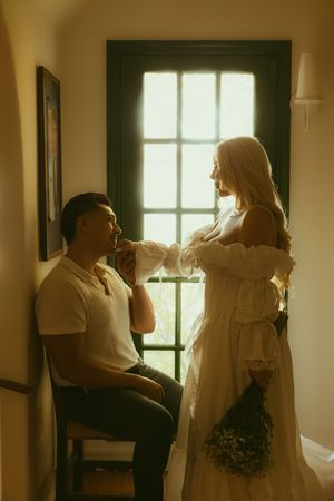 A man seated by a window kisses a woman's hand as she stands in a white off-shoulder dress holding a bouquet.