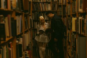 Two people browse books together in a cozy, dimly lit library aisle surrounded by shelves of books.