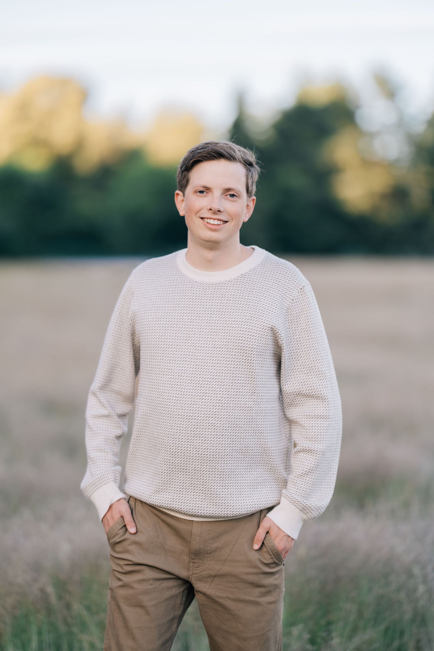 Young man in cream waffle knit sweater smiling outdoors in golden field with trees in background.