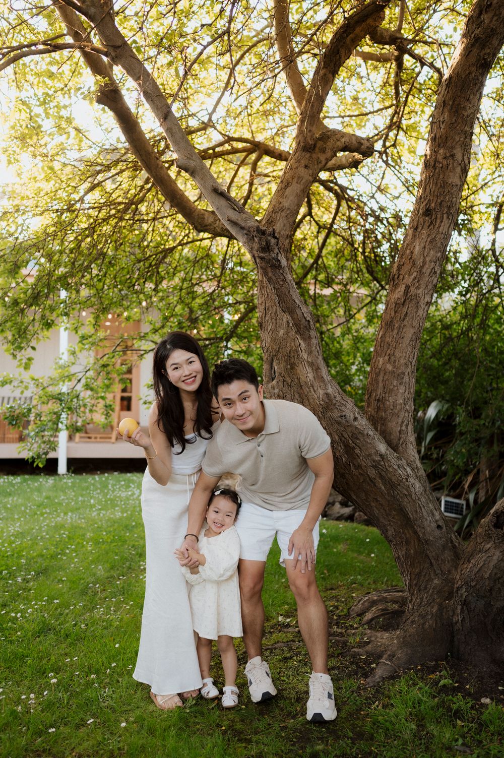 Happy Asian family of three posing under a large tree in a lush green garden on a sunny day.