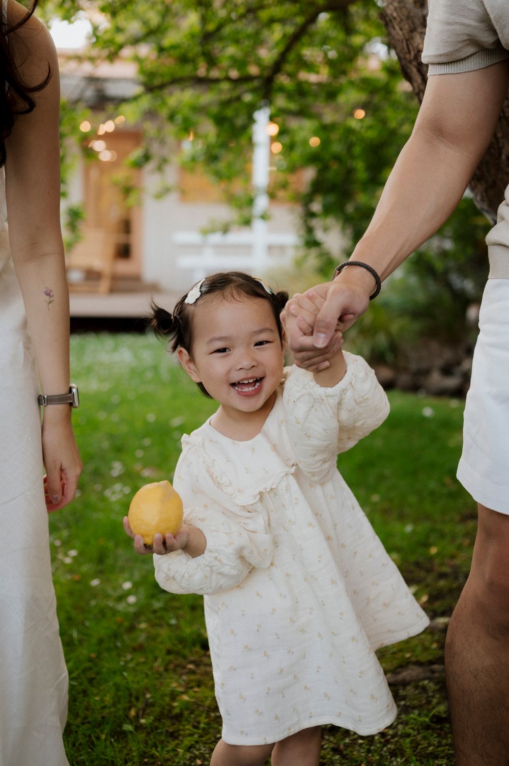 Smiling toddler girl in white dress holding a lemon while standing between parents in a garden.