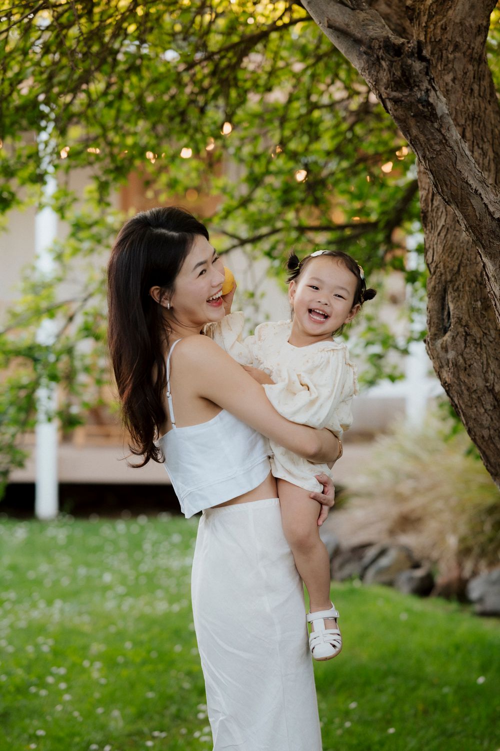 A young Asian woman in white holds a laughing toddler girl under a tree in a lush green garden.