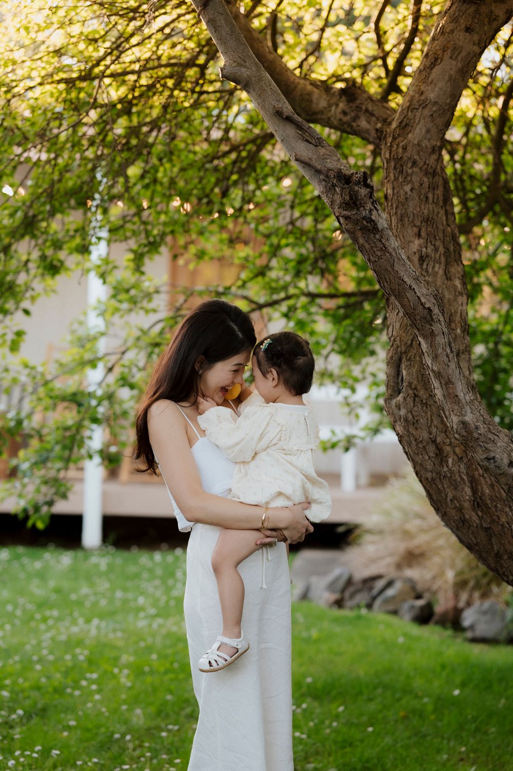 A mother in a white dress tenderly holds her baby girl under a lush green tree in a sunlit garden.