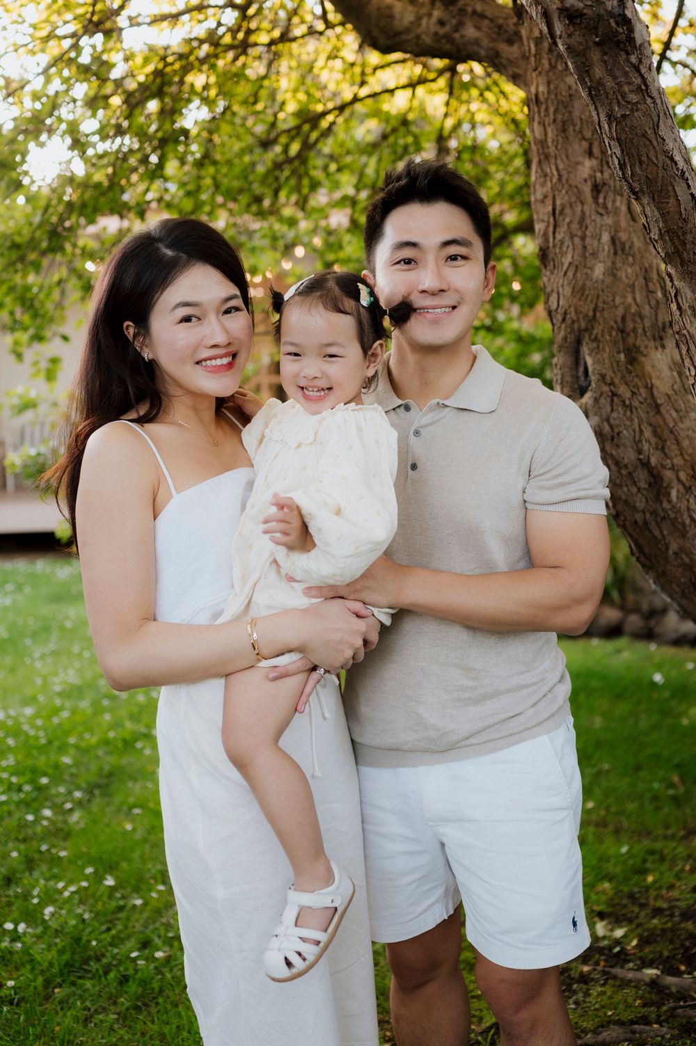 A happy Asian family of three poses outdoors near a tree, parents holding their smiling toddler daughter.