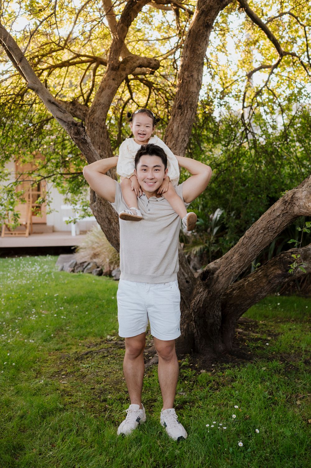 Man carrying smiling child on shoulders in lush garden with large tree backdrop on sunny day.