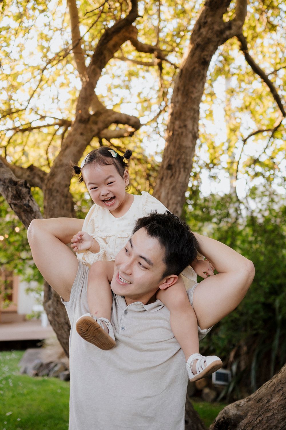 A smiling man carries a laughing toddler girl on his shoulders outdoors beneath a large tree with golden leaves.