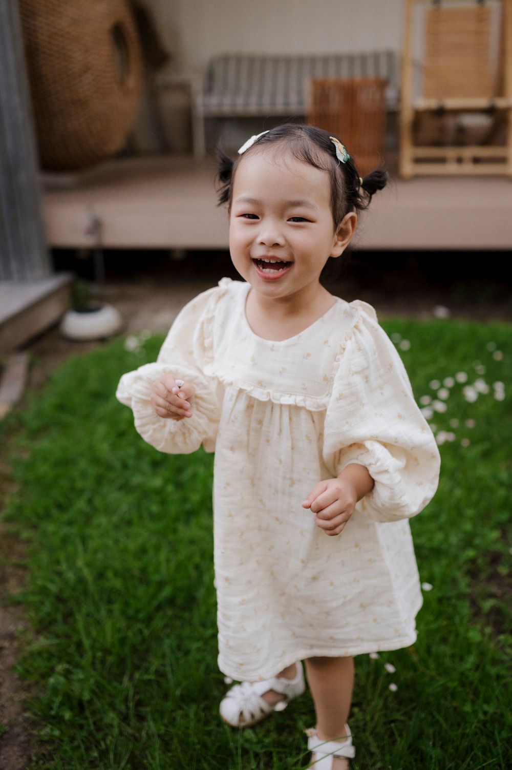 Smiling toddler girl in cream dress with pigtails playing outdoors on green grass near a house.