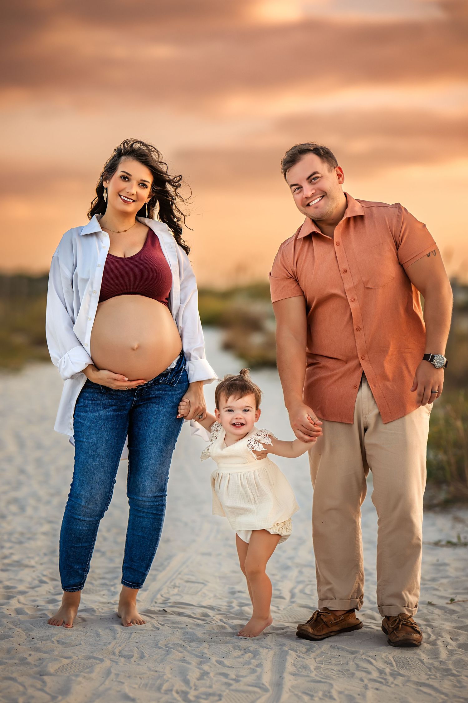Pregnant woman and man swing toddler between them on sandy beach path at golden sunset.