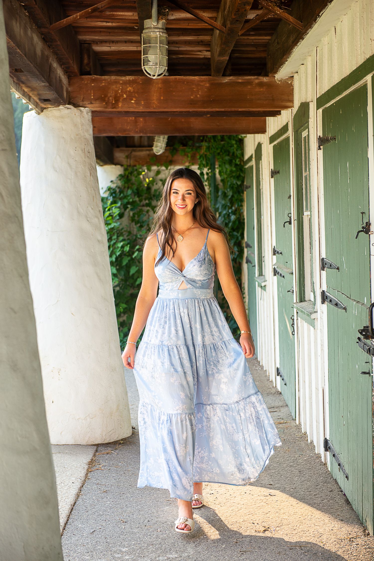 Young woman in a light blue floral maxi dress walking along a rustic covered pathway with green doors.