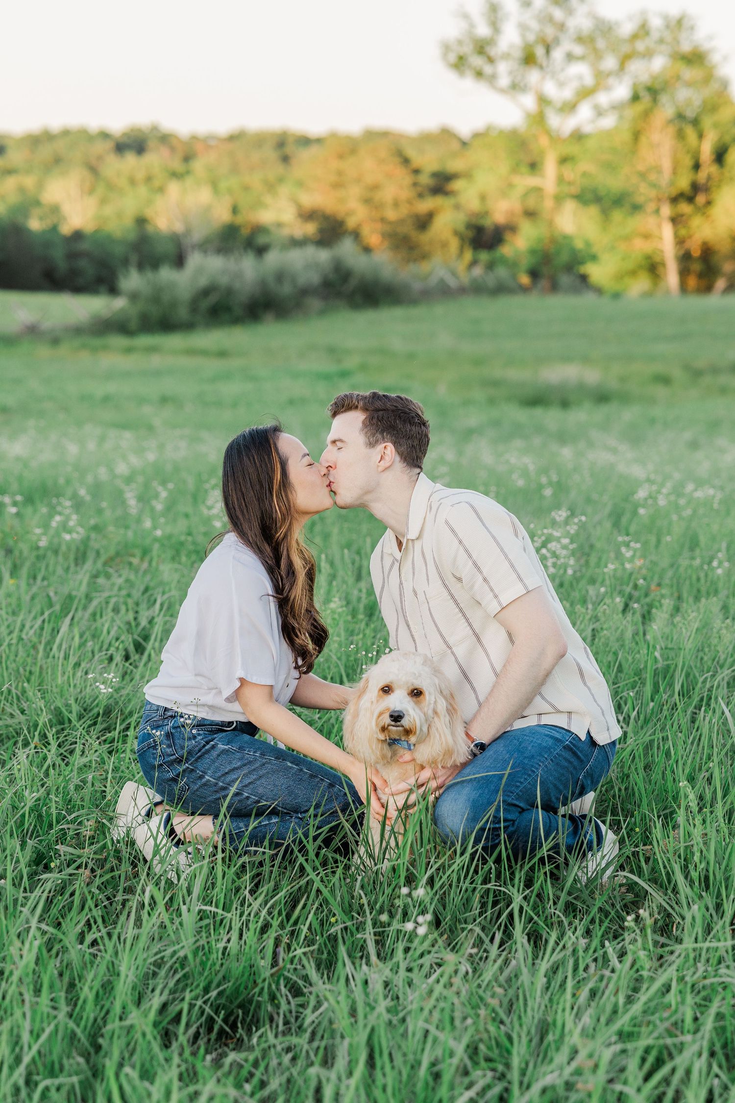 Couple kissing in a green field while holding a small golden dog during a romantic outdoor engagement photo session.