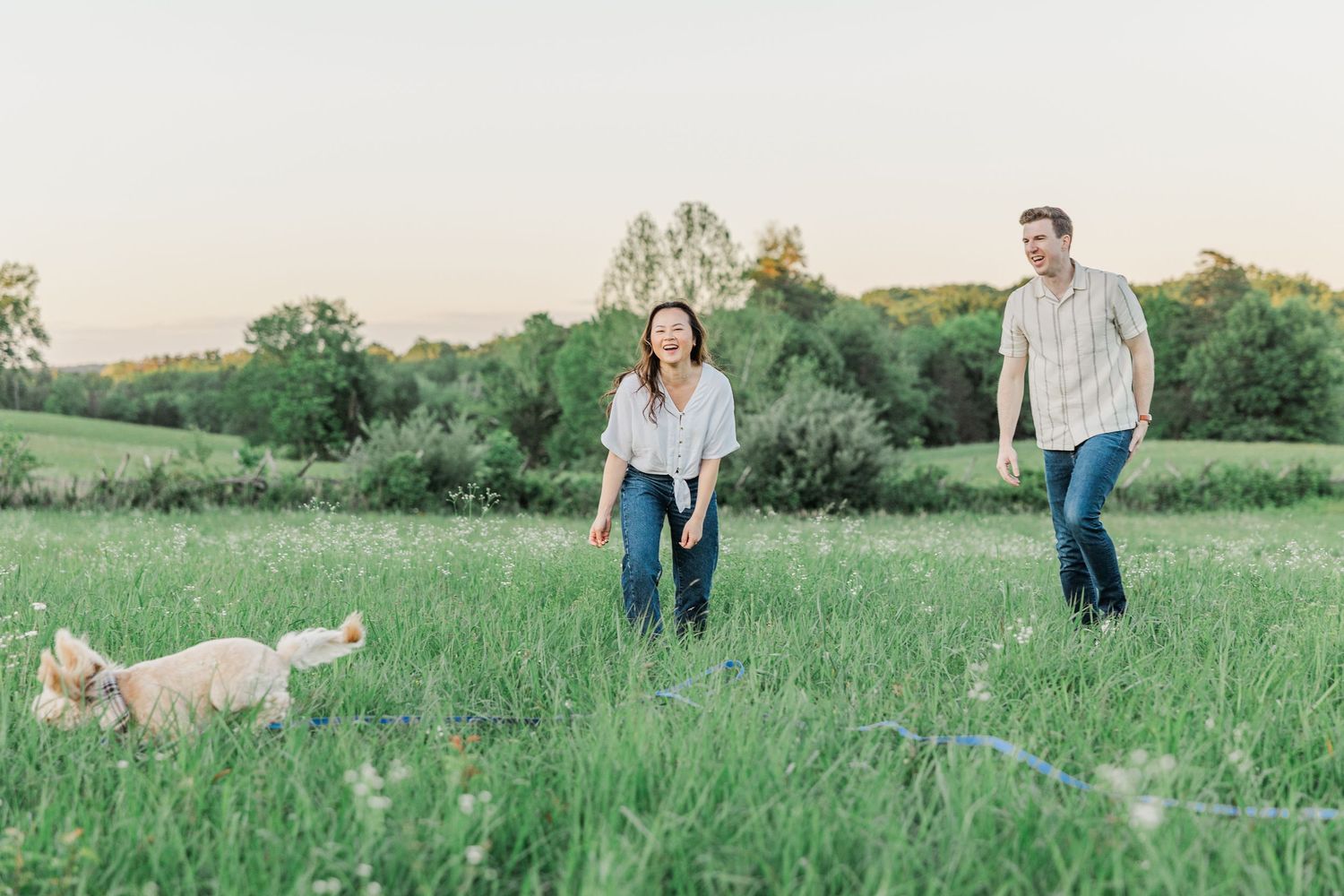 Couple laughing with golden retriever dog in lush green meadow at sunset.