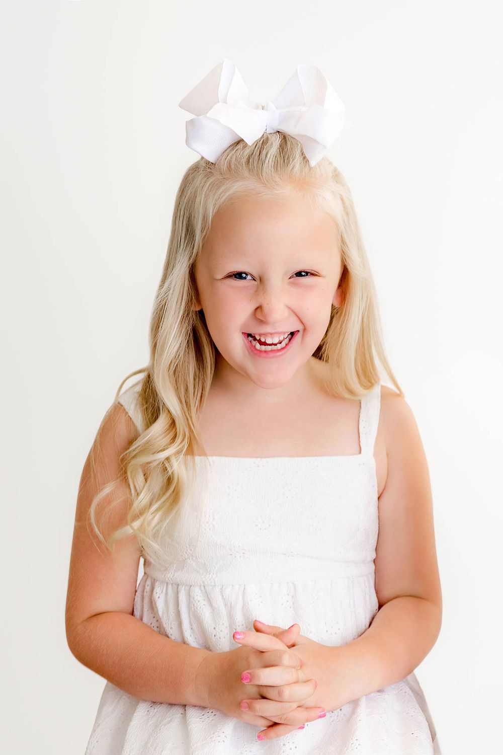 Smiling blonde girl in white dress with large white bow hair accessory on white background.