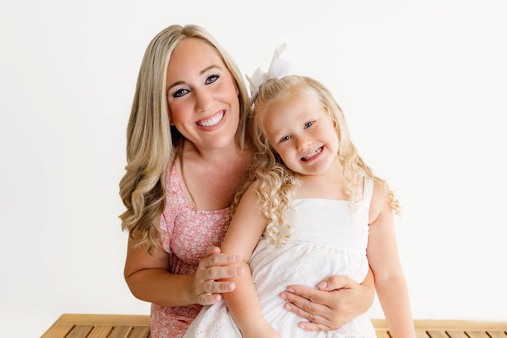 A smiling blonde woman in a floral dress poses with a curly-haired little girl in white on a bench.
