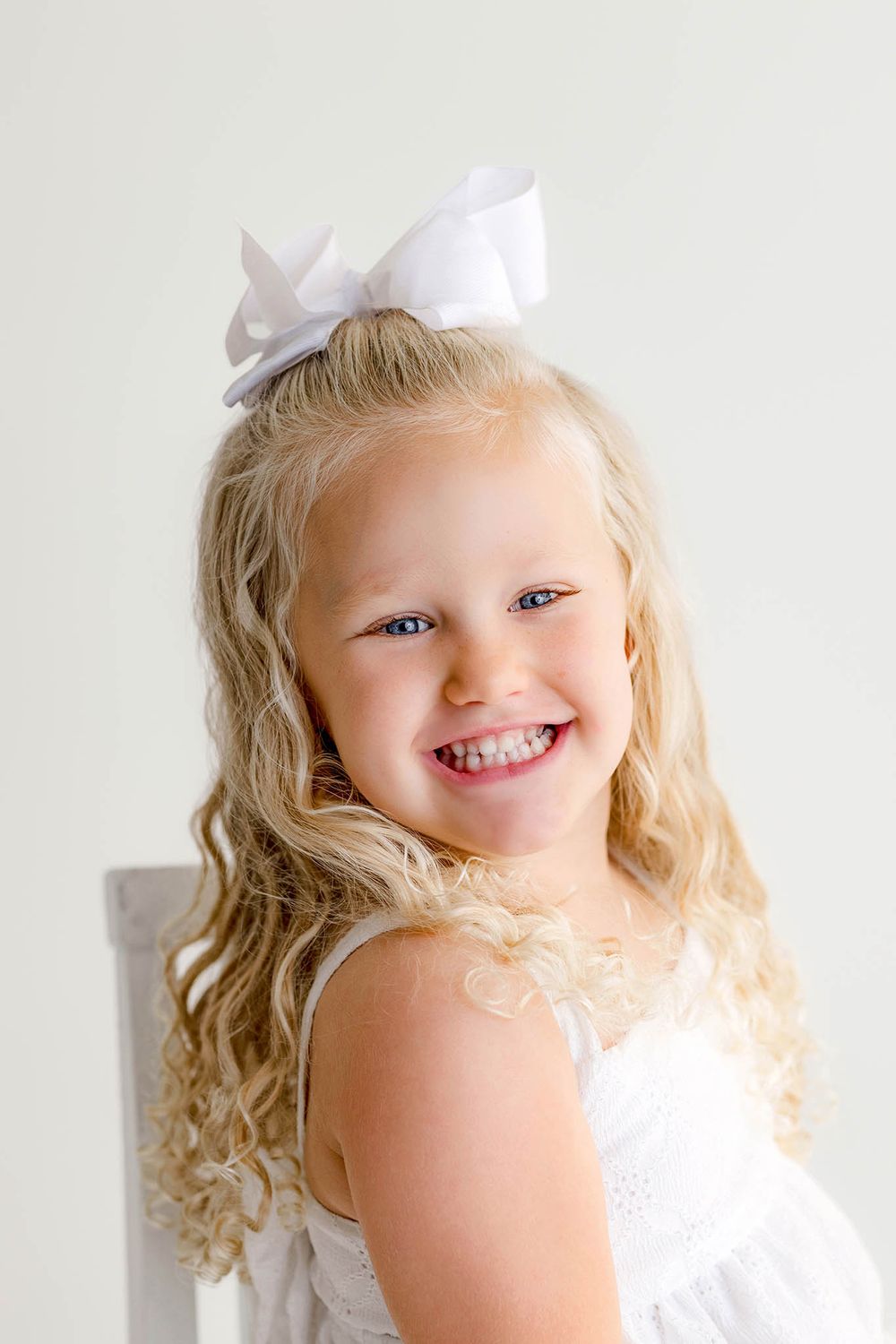 A smiling blonde girl in a white dress with a bow poses for a portrait photo by Temecula photographer.