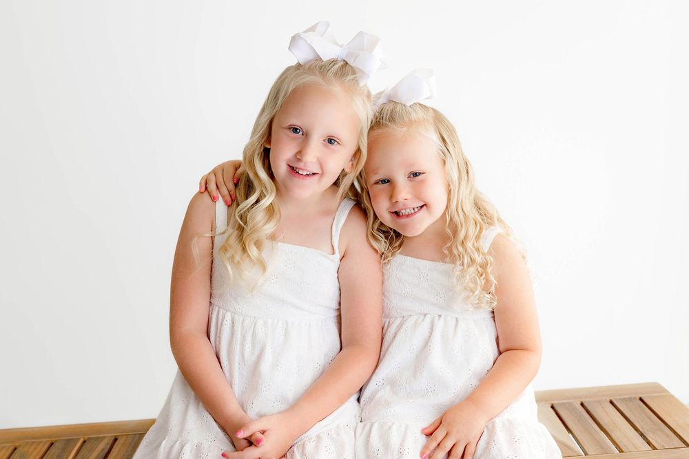 Two smiling blonde girls in matching white dresses and bow hairpieces sitting together on a bench.