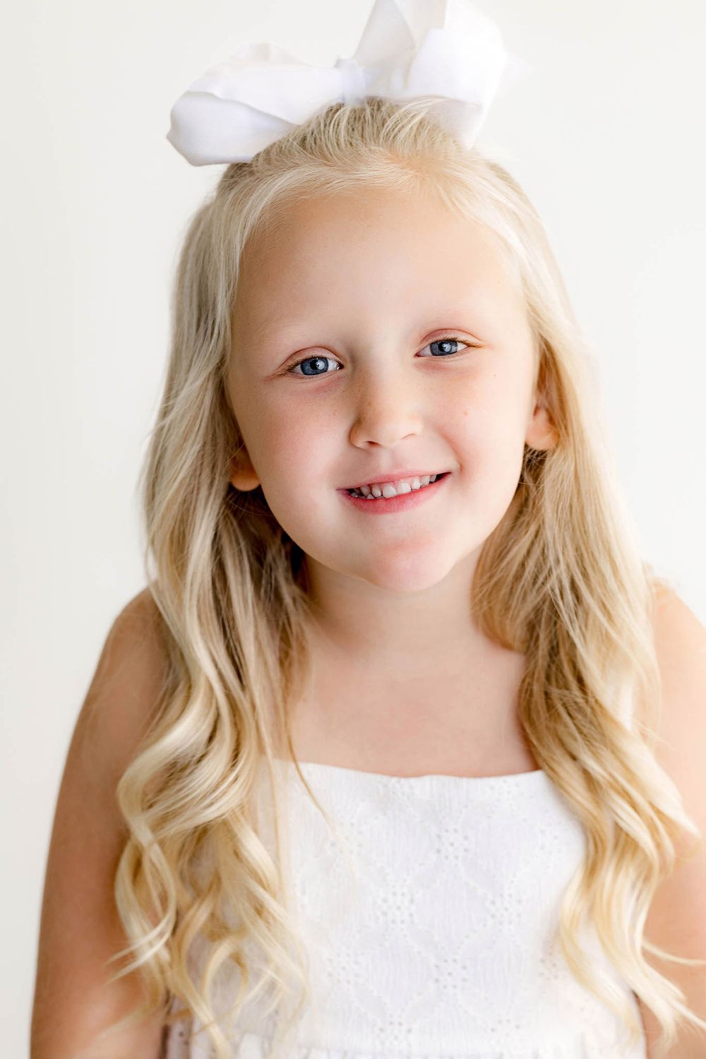 Smiling blonde girl with long curls wearing white bow and white eyelet dress.