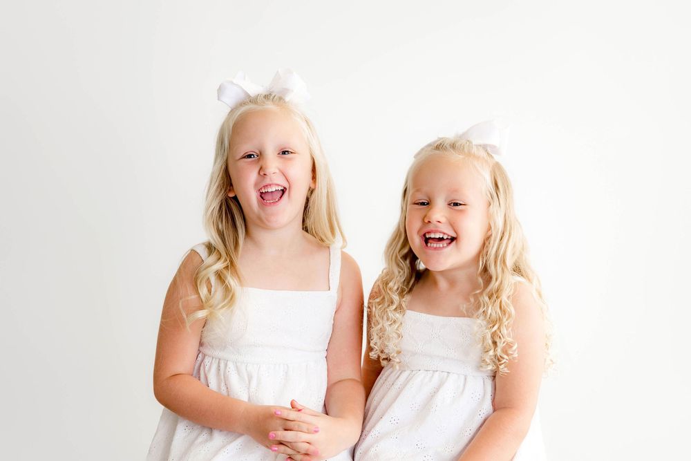Two laughing blonde girls in matching white dresses with white bows pose together against a white background.