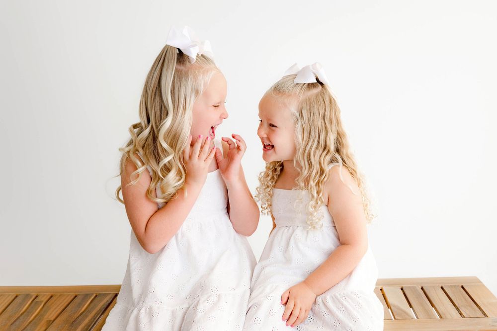 Two young blonde girls in white dresses with bow hair clips laugh together on a wooden bench by Temecula photographer.