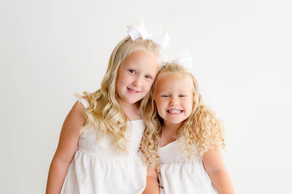Two smiling blonde girls in matching white dresses with bow hairpieces pose together on white background by Temecula photographer.