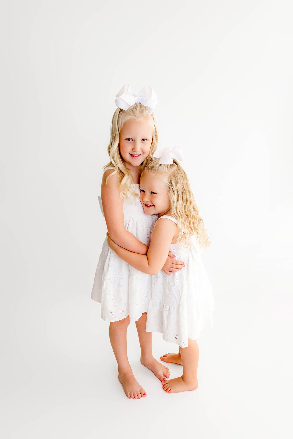 Two young blonde sisters in white dresses and bows share a sweet hug against a white background.