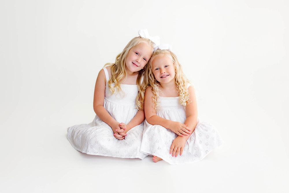 Two young girls in matching white dresses sitting together and smiling on a white background by Temecula photographer.