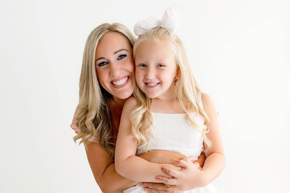 A smiling blonde woman hugs a young girl with a white bow in her hair against a white background.
