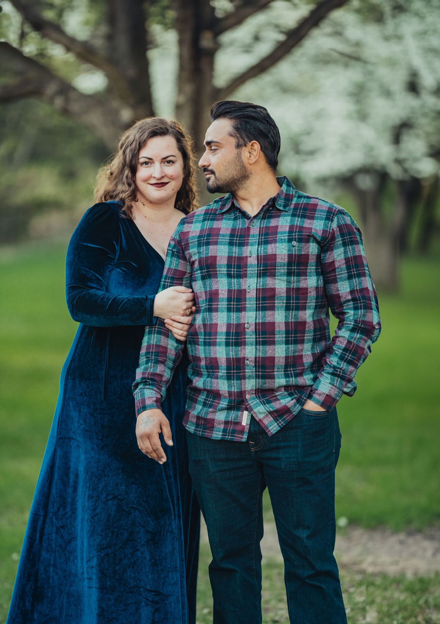 Couple posing outdoors in a lush green park, woman in blue velvet dress and man in plaid shirt smiling together.