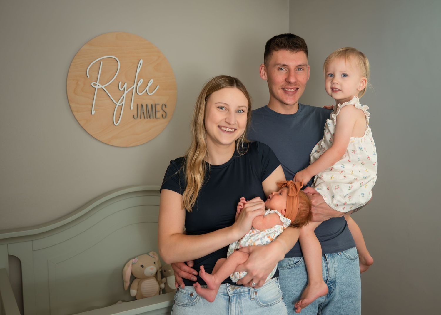Happy family of four posing in nursery, parents holding newborn and toddler near Rylee James name sign.